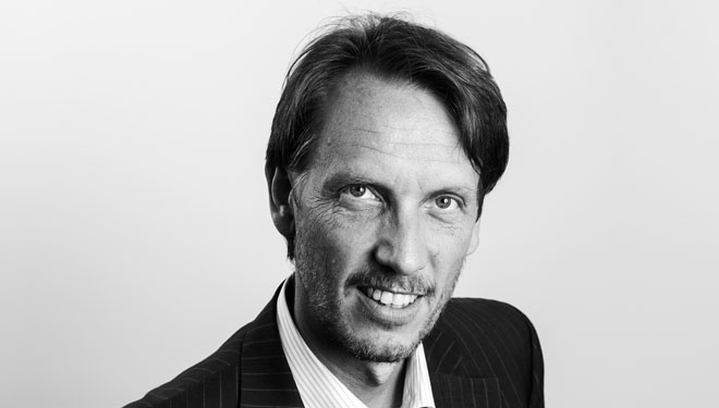 Professional headshot of a smiling man in a dark pinstripe suit against a light backdrop
