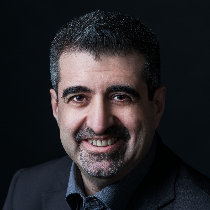 Close-up portrait of a smiling man with dark hair and a goatee, wearing a dark blazer against a black background.